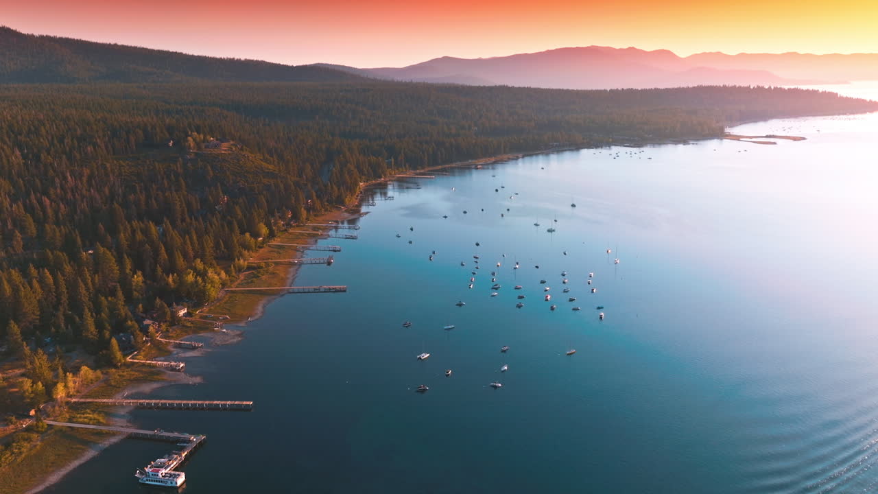 Wooded shore of majestic Lake Tahoe at sunset. Numerous boats sailing on the water. Pink skies and mountains at backdrop.