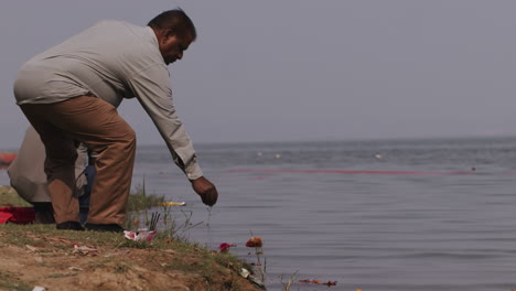 a hand held long shot of an indian man sitting with his back towards the camera on the banks of a river and doing the last rites, as per hindu religion, of a dead person by throwing ashes in water