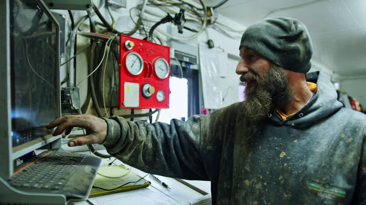Charismatic bearded male wearing a cap and dirty clothes stands at the computing equipment of oil production site. Man touches the screen operating a machine.
