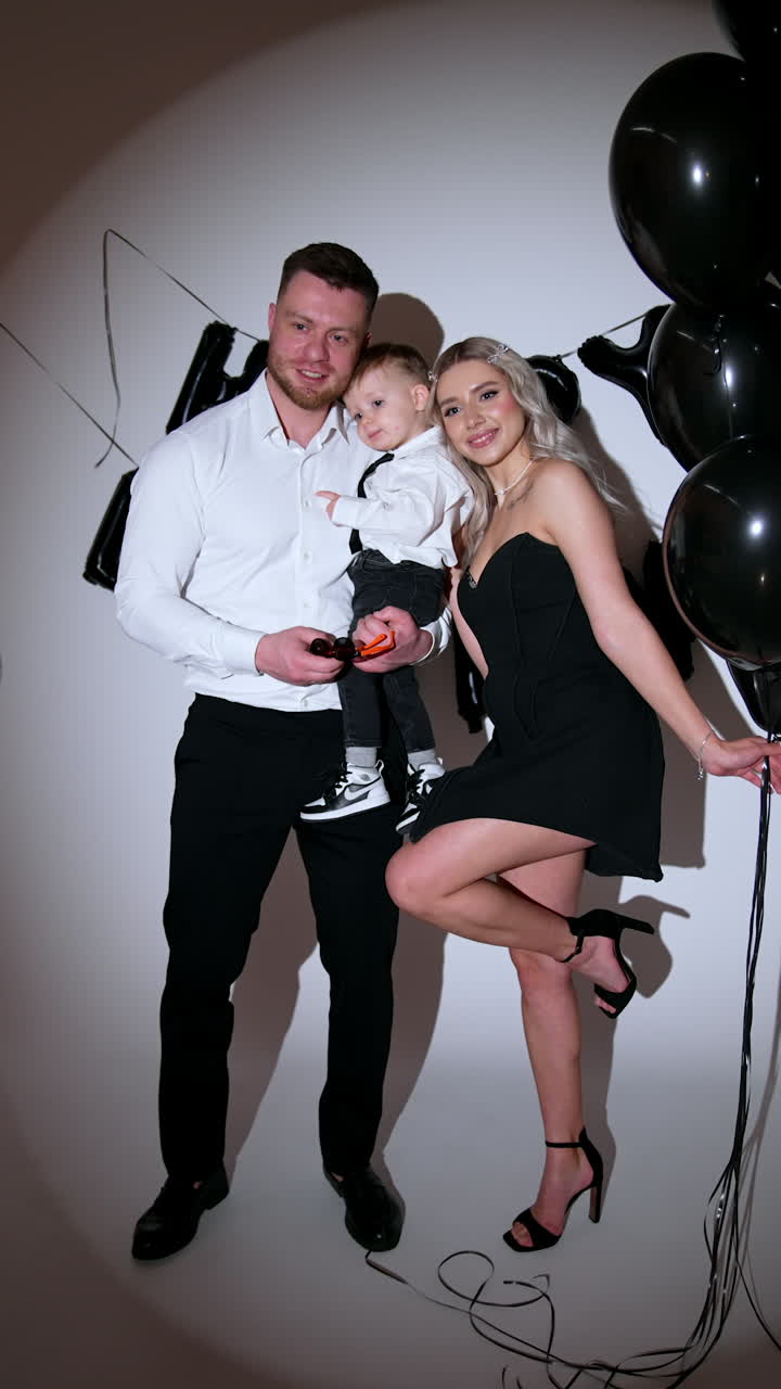 Family of three wearing black and white clothes stand in studio. Woman is holding a bunch of black balloons. Vertical video.