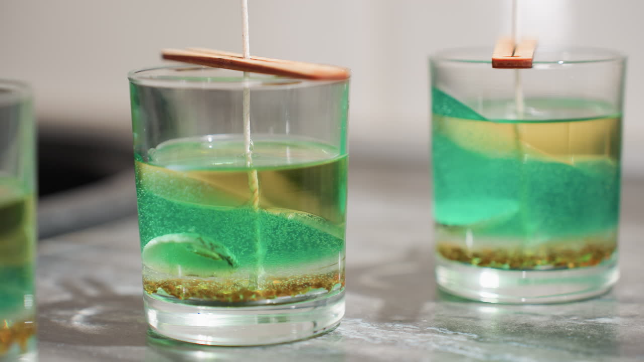 Close up of tealight cup filled with layered green and brown wax mixture with embedded particles, wooden sticks placed on top holding centered wick straight inside container