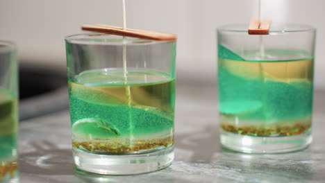 close up of tealight cup filled with layered green and brown wax mixture with embedded particles, wooden sticks placed on top holding centered wick straight inside container