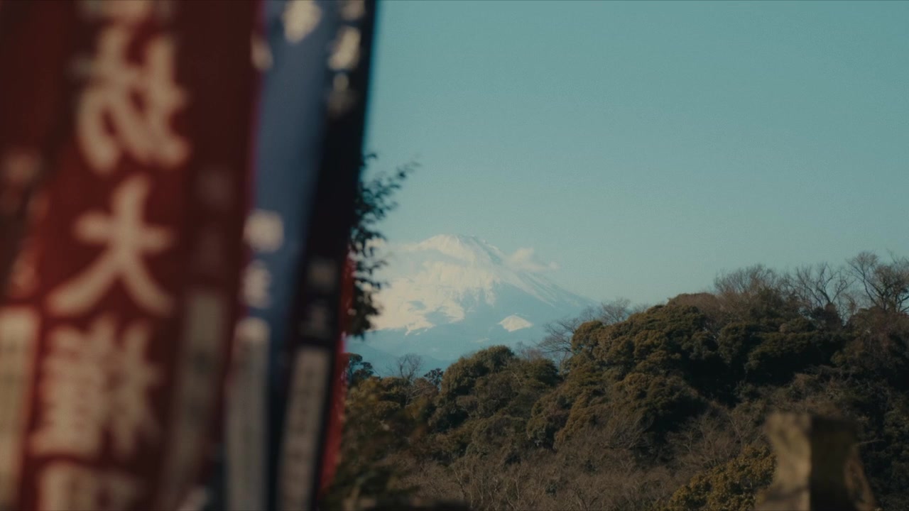 A distant view of snow-capped Mount Fuji seen from the Hansobo Shrine observation deck atop Kencho-ji temple in Kamakura, with traditional red and white shrine banners in the foreground.