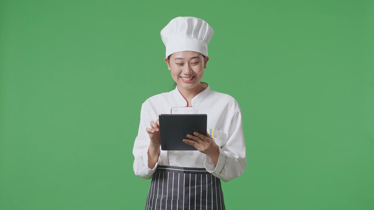 Asian Woman Chef Smiling And Pointing To Tablet In Her Hand While Standing In The Green Screen Background Studio