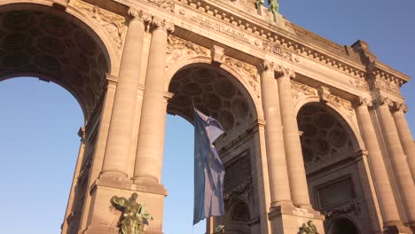 the eu flag sways beneath the monumental cinquantenaire arch in brussels, belgium, with the neoclassical columns and coffered ceilings bathed in soft golden light during a clear afternoon