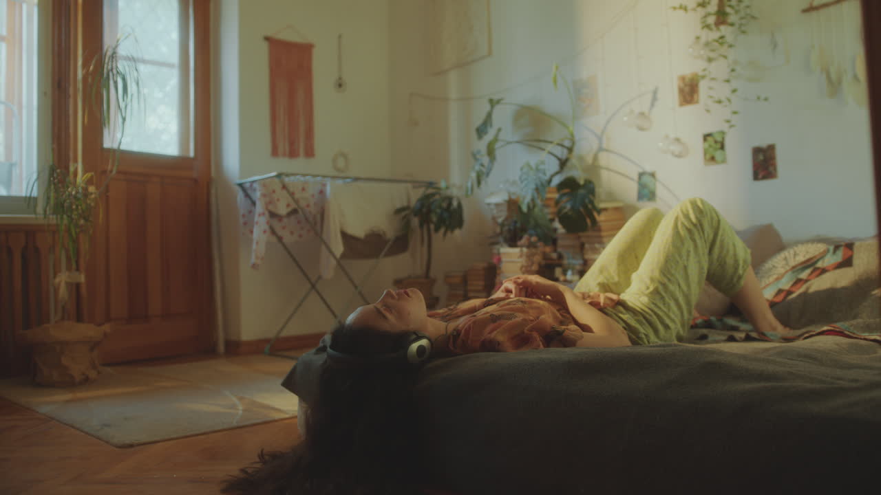 젊은 여성이 헤드폰으로 음악을 듣고 빛 방에서 침대에서 휴식을 취하고 있습니다.