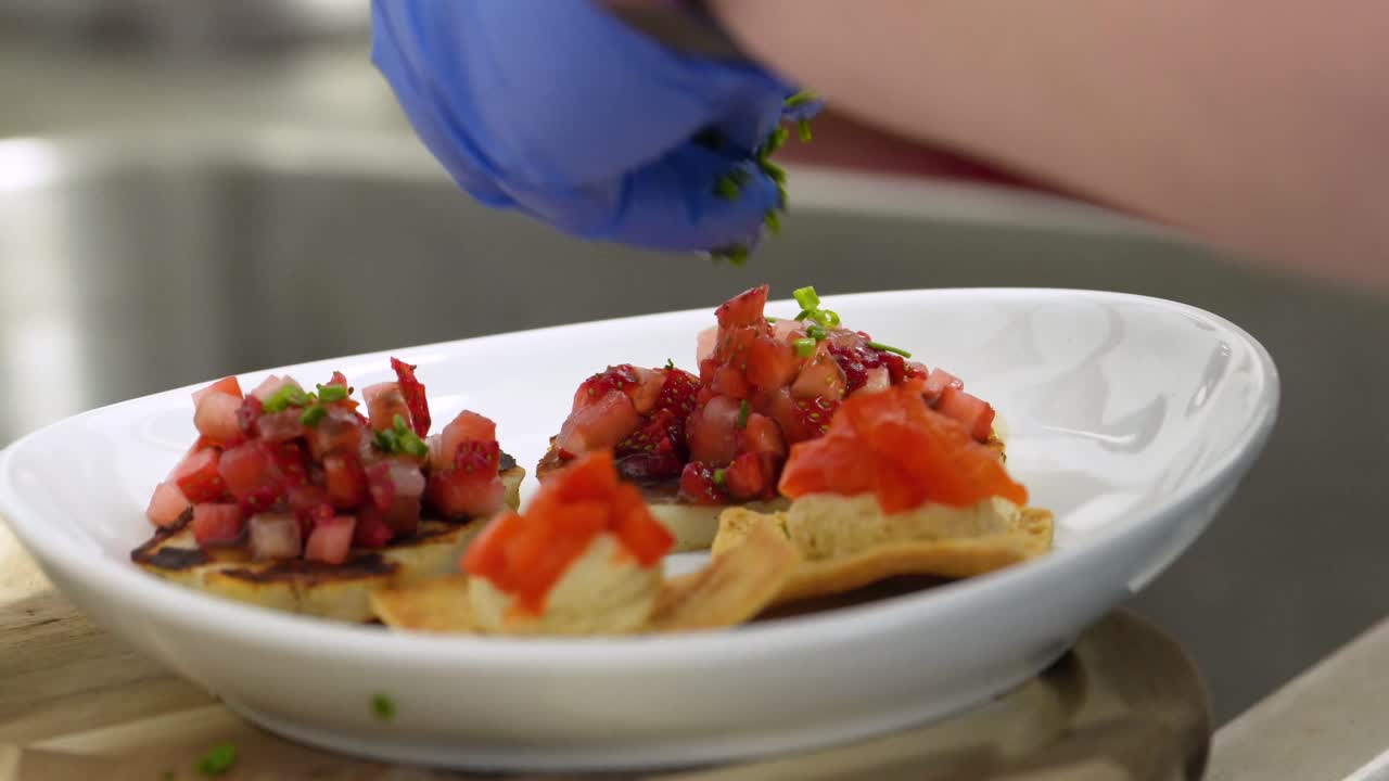 Cook Wearing Blue Rubber Gloves Seasons Healthy Appetizer Meal with Herbs on Diced Tomatoes and Pita Bread Chips with Hummus, in the Kitchen using a White Bowl or Plates, Food Closeup