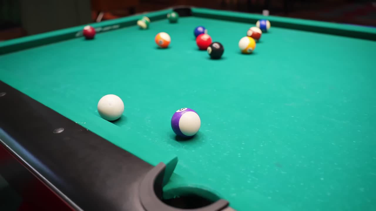 Speelbal en paarse gestreepte bal liggen bij de pocket op een groene pooltafel, met andere ballen wazig op de achtergrond onder gezellige barverlichting en ondiepe scherptediepte.