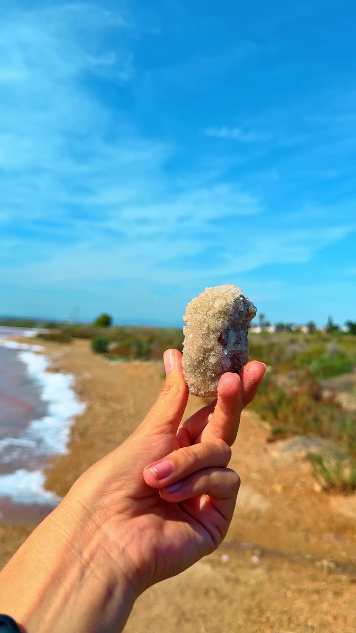 Mano che tiene un grande cristallo di sale su uno sfondo lacustre. Primo piano di una mano umana che tiene un cristallo minerale di sale grezzo con un lago rosa e la riva sullo sfondo.