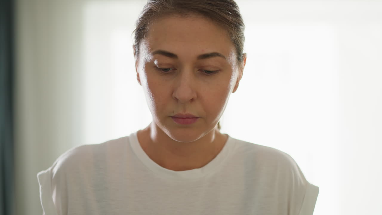 mujer pensativa mira hacia abajo en un primer plano de fondo ligero. concentrada dama en blusa blanca de pie por la ventana en la habitación. retrato de mujer bonita en el estudio