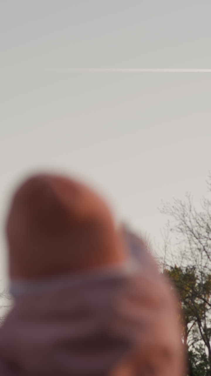 A man in a green jacket carries his daughter, dressed in a pink jacket and matching beanie, as they both point towards an airplane streaking across the sky