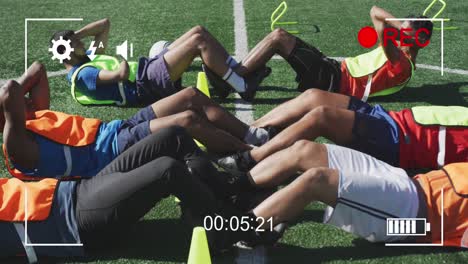 Video-camera-digital-interface-against-team-of-male-soccer-players-performing-crunches-exercise