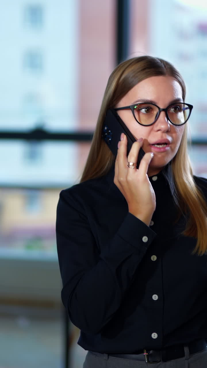 Young woman speaking on the phone and walking in the office. Emotional conversation over the phone. Couple at the table at backdrop. Vertical video