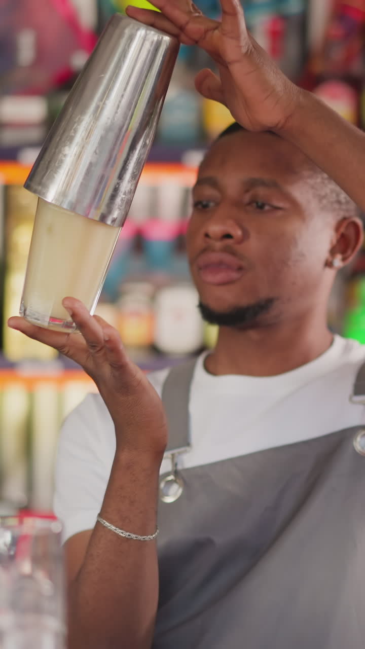 Focused bartender shakes cocktail in metal cup in bar. African American barman scrupulously prepares drink mixing ingredients in shaker. Black man works in bar