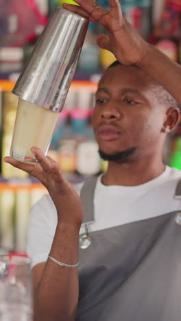 el camarero enfocado agita el cóctel en una taza de metal en el bar. el camarero afroamericano prepara escrupulosamente la bebida mezclando ingredientes en el agitador. el hombre negro trabaja en el bar
