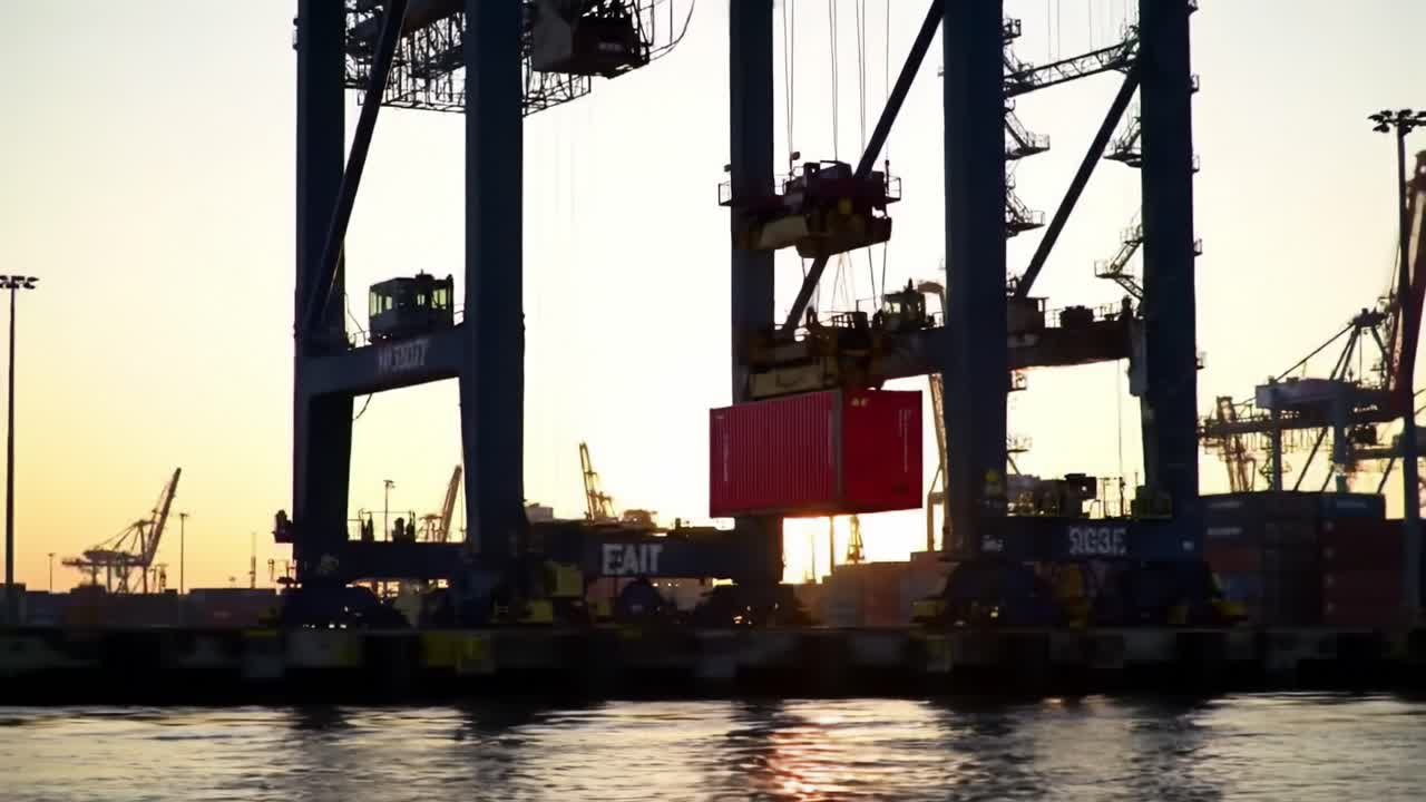 Cranes operate to lift and load large containers onto ships in a bustling port during a vibrant sunset. The scene captures the dynamic nature of maritime logistics and trade.