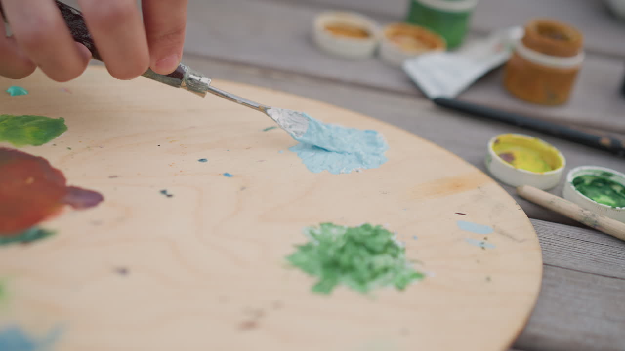 palette da strada con pigmento blu mescolato su una tavola, la mano di un muralista usa un coltello di metallo per preparare una spessa pasta acrilica, barattoli e tubetti dai colori vivaci sparsi su un tavolo di legno, bokeh superficiale, luce esterna dinamica