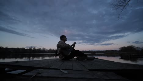 silueta de músico en negro tocando la guitarra sentado en el terraplén del muelle al atardecer