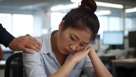 a young woman appears distressed at her workstation in a professional environment. she leans her head on her hands, suggesting feelings of fatigue and pressure as coworkers work nearby.