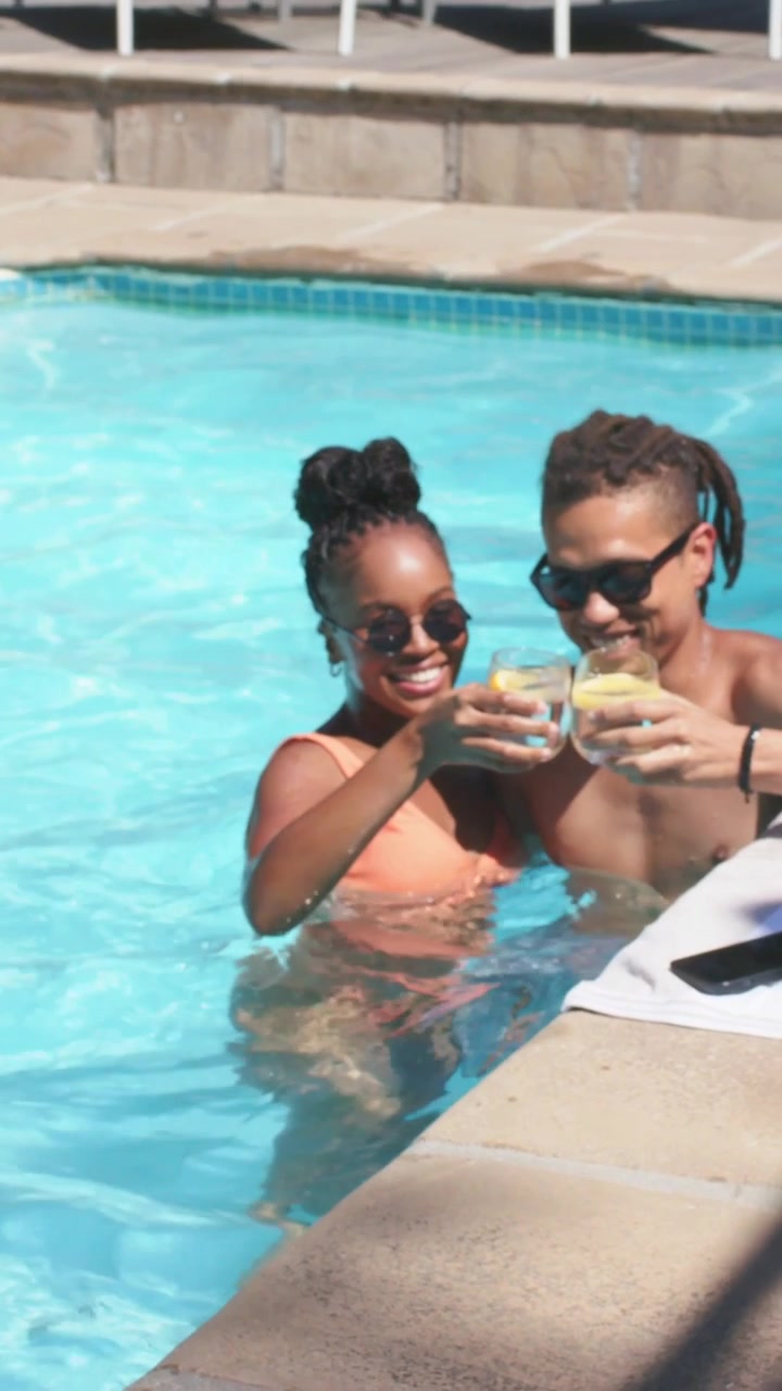 Vertical video: Hugging woman in swimsuit reaching for drink on edge, giving glass to man at pool