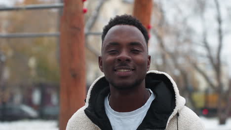 smiling african man wearing warm winter jacket standing outdoors with snow-covered ground and bare trees in background, his cheerful expression contrasts with cold weather