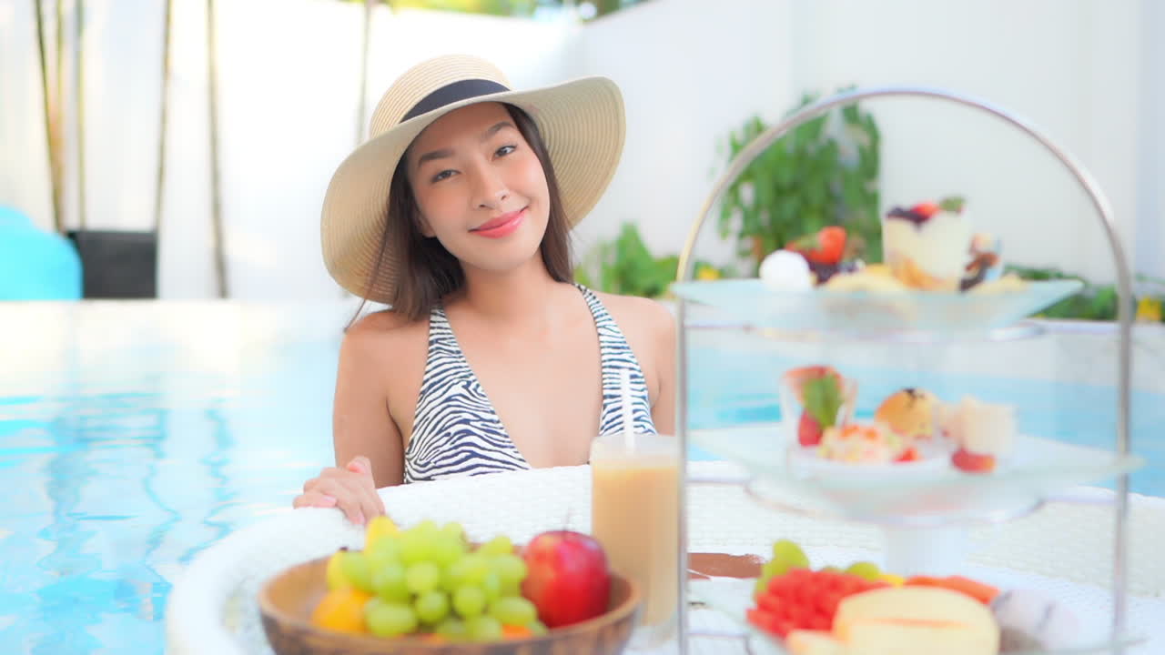 Fit young Asian woman with floating exotic breakfast tray full of fruits and deserts in a swimming pool in the luxury resort, close up