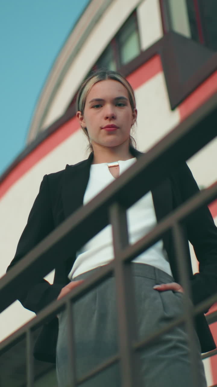 Upward view of confident lady posing with warm smile near iron railing with striped office building in background under clear blue sky, hands in pockets and radiating poise