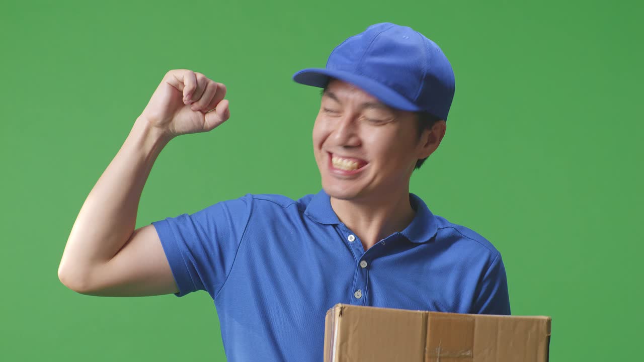 푸른 유니폼을 입은 아시아 남성 택배사의 클로즈업 미소와 근육을 구부리는 동안 스튜디오에서 녹색 화면 배경에 상자를 배달