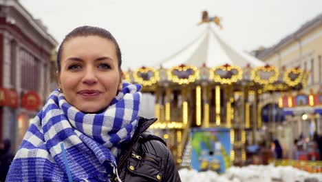 beautiful smiling brunette in warm scarf looks at camera at carousel background