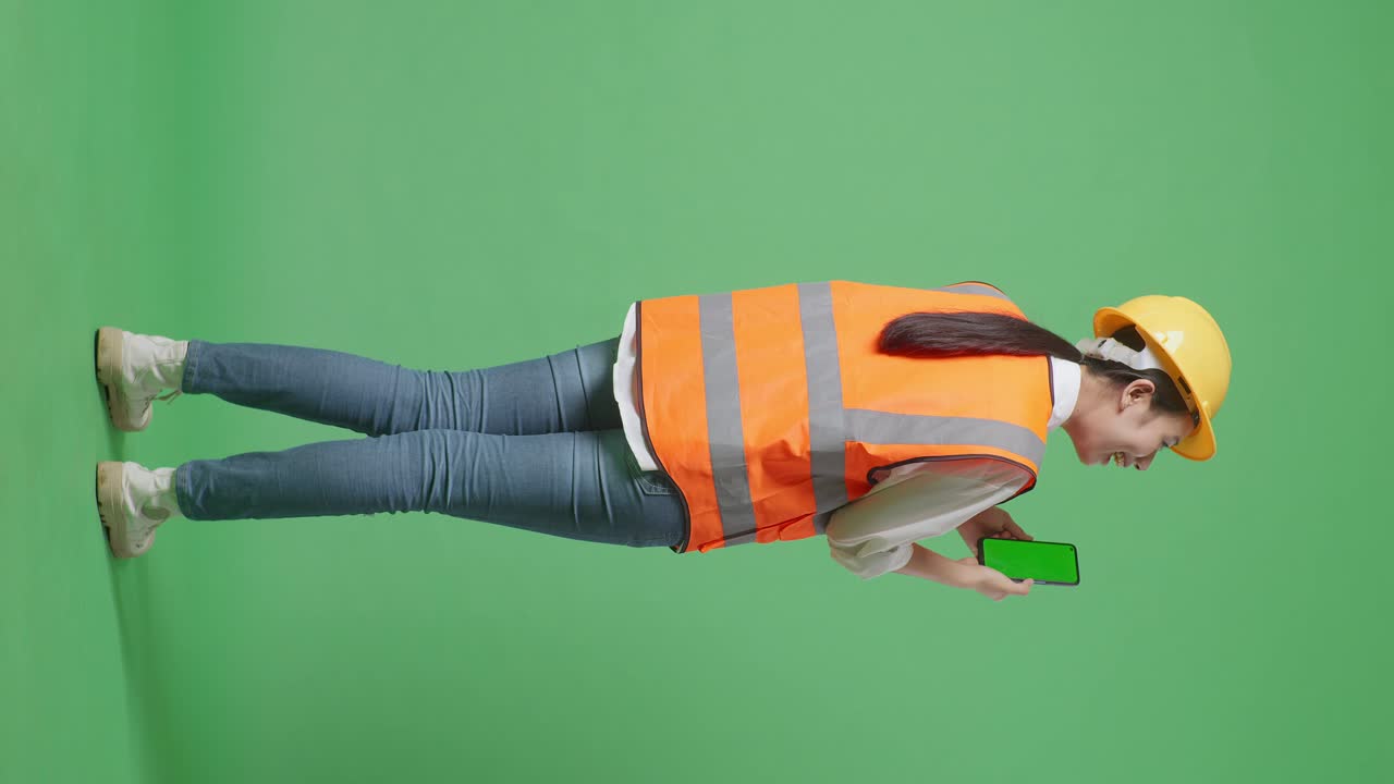Full Body Back View Of Asian Female Engineer With Safety Helmet Working On A Green Screen Smartphone And Looking Around While Standing In The Green Screen Background Studio