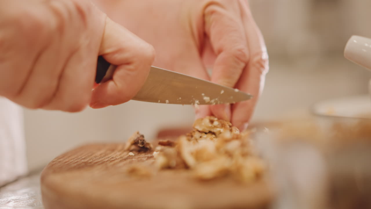 Primo piano di mani che affettano noci su un tagliere, con il movimento del coltello. Luce calda della cucina, preparazione di un topping croccante per la torta di zucca. Profondità di campo ridotta, accogliente scena di cottura autunnale, texture rustica e preparazione accurata.