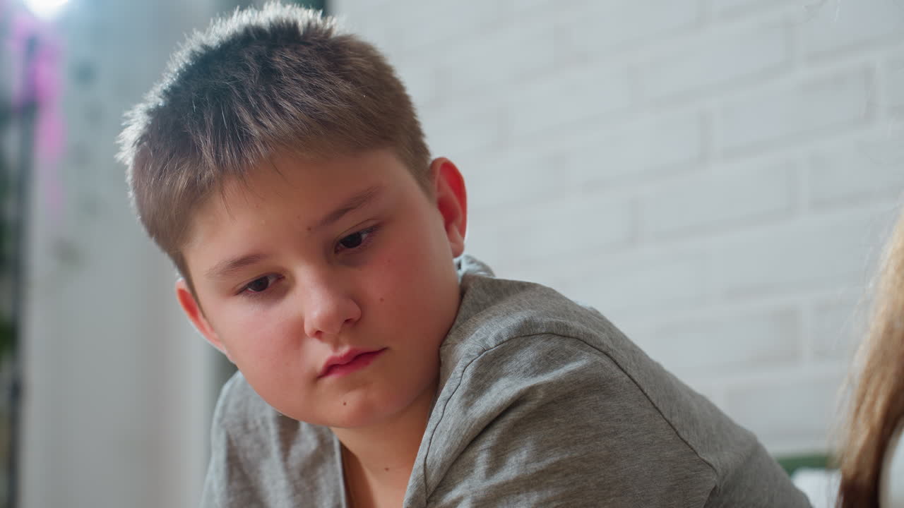close view of boy in grey top looking serious and focused while observing book page, soft indoor lighting with white brick background, expression of deep concentration and curiosity