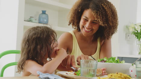 Madre-Ayudando-A-Su-Hija-A-Cortar-Comida-Sentada-Alrededor-De-La-Mesa-En-La-Cocina-De-Casa-Comiendo-Juntos