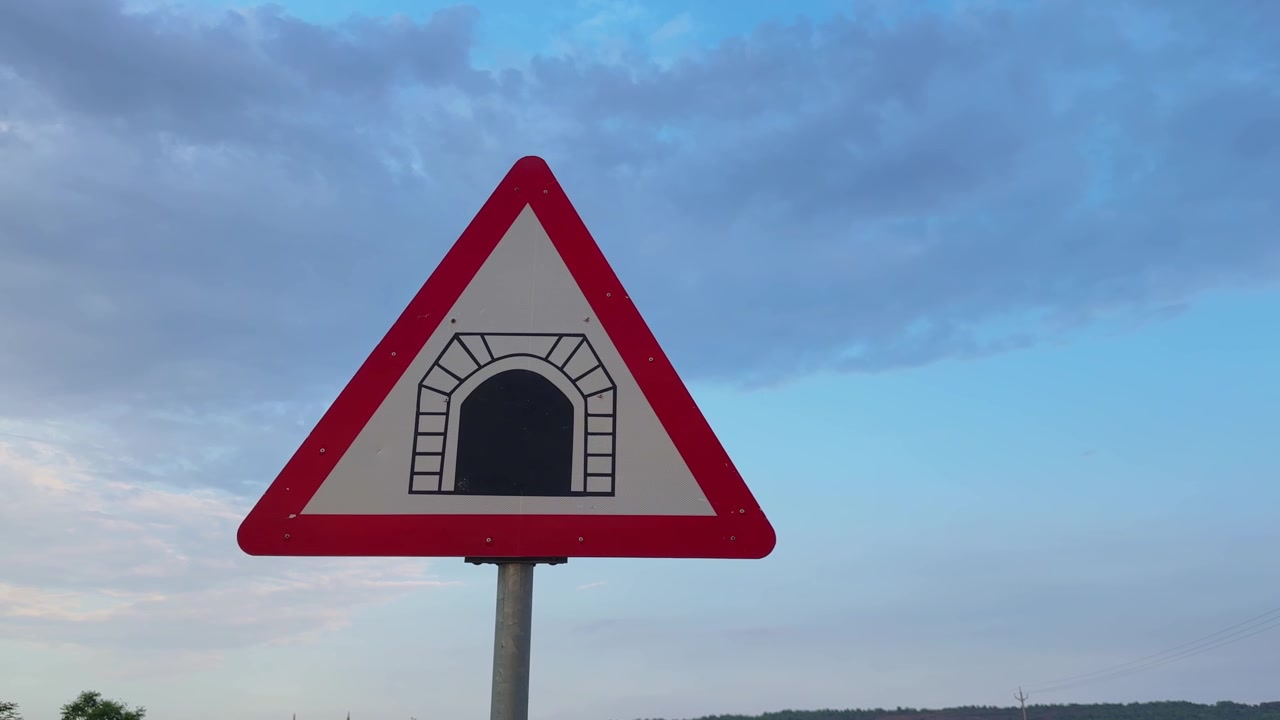 Camera slowly orbiting around a triangular tunnel warning road sign against a dramatic cloudy sky, highlighting transportation safety, traffic guidance and roadside infrastructure.