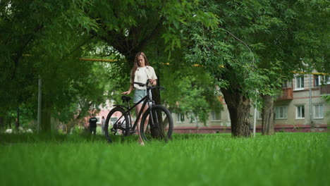 young woman holds black gloves as she approaches her parked bicycle on a paved path, preparing to wear gloves amidst lush greenery, background features trees, people passing by