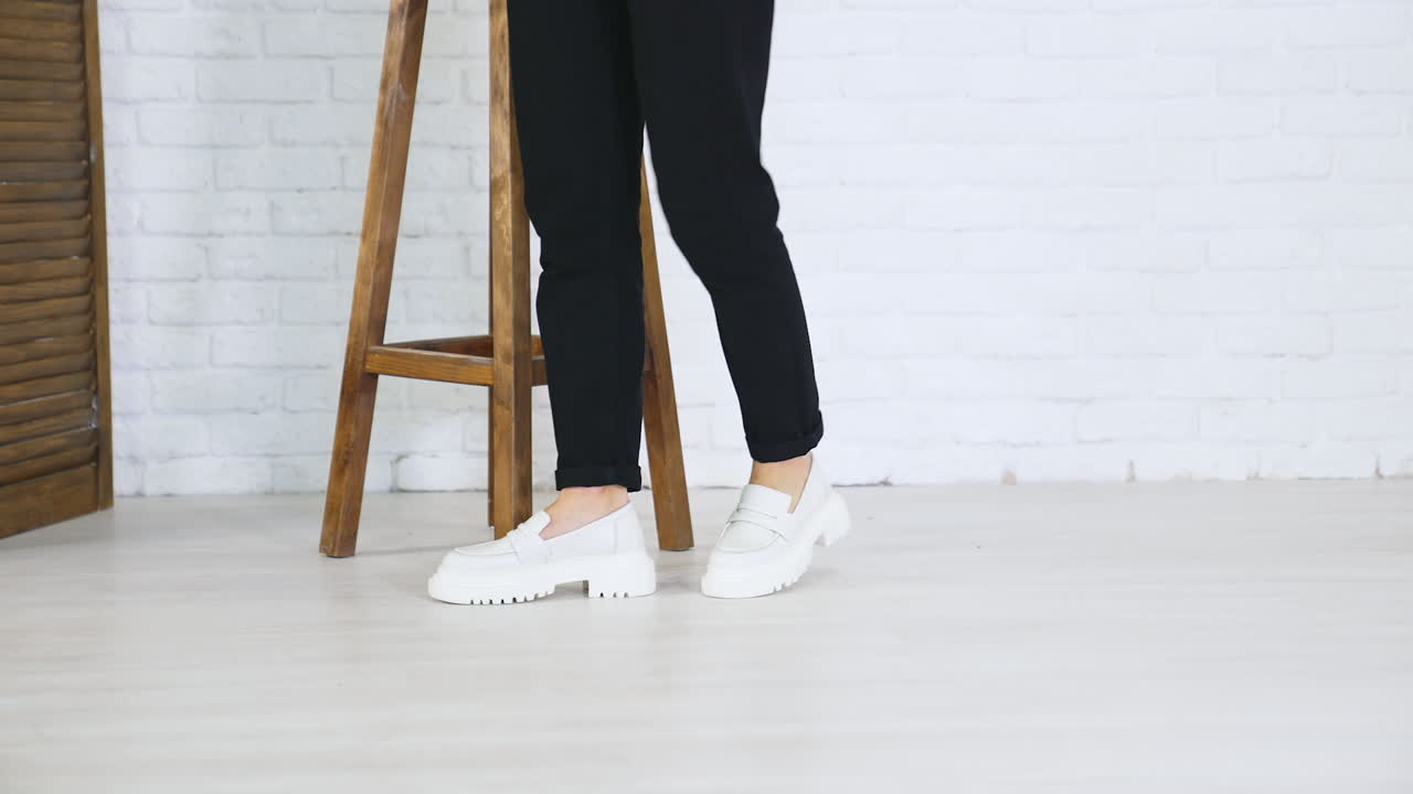 Fashionable white shoes presentation. Female model walks in fashionable footwear back and forth near the wooden tabouret.