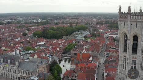 Passing-Belfry-of-Bruges-Belltower-revealing-Cityscape-and-River,-Aerial-forward