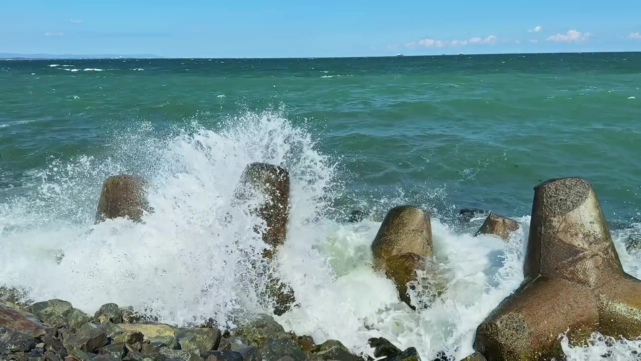 Le onde si infrangono contro barriere di cemento in riva al mare durante una giornata di sole in una zona costiera con cielo blu e barche visibili in lontananza