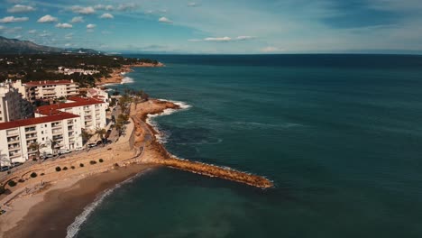 Zoomen-Sie-Aus-Der-Küstenpromenade-Von-Ametlla-De-Mar-In-Tarragona,-Spanien,-Mit-Wellen,-Die-An-Einem-Sonnigen-Tag-Gegen-Die-Küste-Schlagen---Luftaufnahme