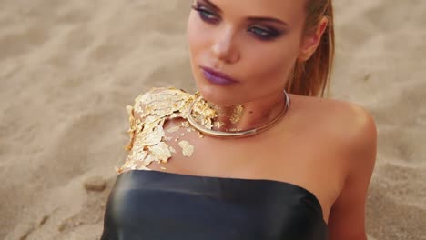 young model with professional golden art makeup in black leather swimsuit lying on the beach looks at the camera. attractive