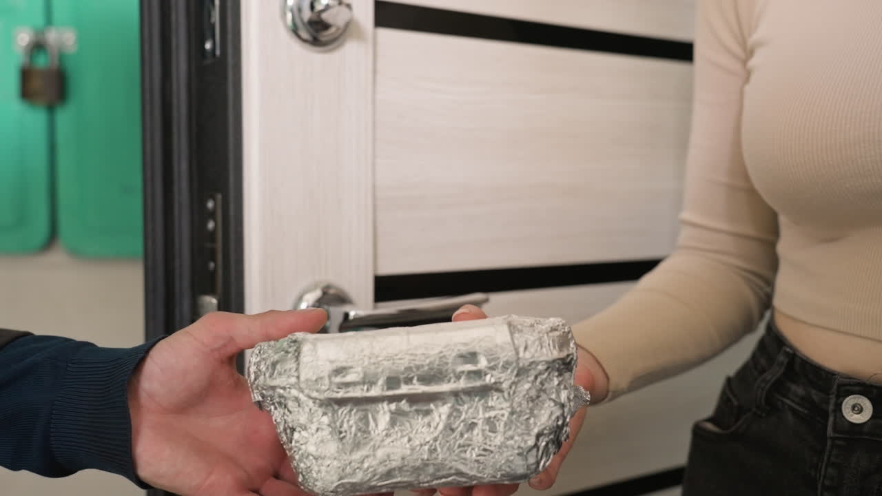 jovem recebe entrega de comida na porta de casa, mãos trocam recipiente embrulhado em papel alumínio, entrega sem contato pelo entregador, moletom bege casual e jeans, interior de apartamento com maçaneta de porta moderna