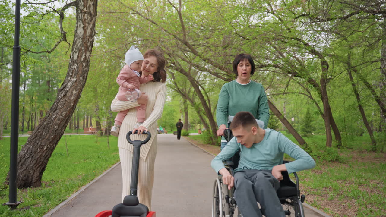 Person Interacts Kindly, Caucasian Male In Wheelchair Shares Helpful Gesture With Baby, Man With White Ethnicity Using Wheelchair Engages Playfully With Child And Woman Observing Nearby