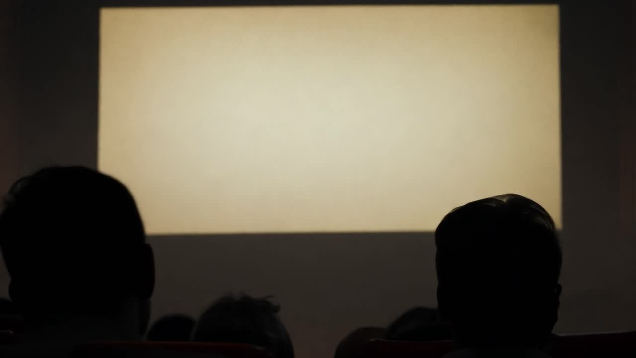 Spectators sitting in a dark theater watch a blank screen, creating an atmosphere of anticipation and curiosity
