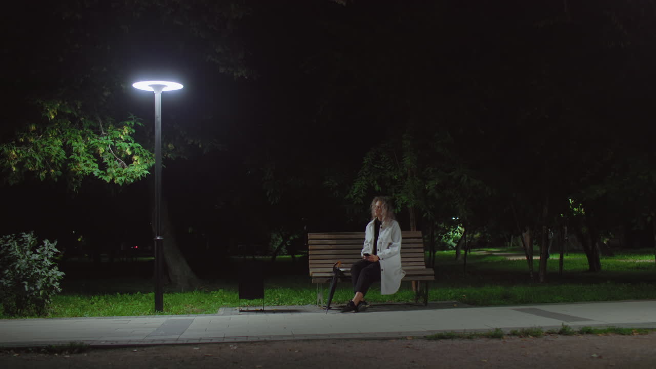 Woman seated alone on park bench under streetlamp at night holding phone in hand while gazing thoughtfully into distant space, surrounded by quiet greenery and soft ambient lighting from nearby lamps