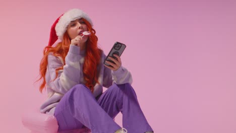 studio shot of young gen z woman wearing christmas santa hat eating candy lollipop looking at mobile phone 3