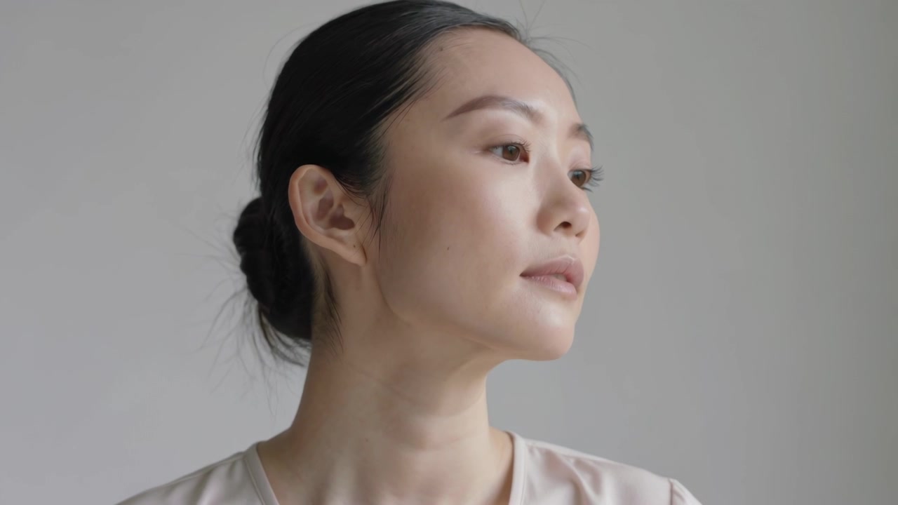スタジオで、明るいブラウスを着用したアジア人女性がゆっくりと頭を右に回し、顎を上げながら回転する。