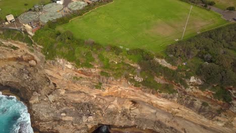 Epic-drone-shot-of-ocean-waves-smashing-against-cliff-and-rocks,-near-a-park,-Sydney,-Australia