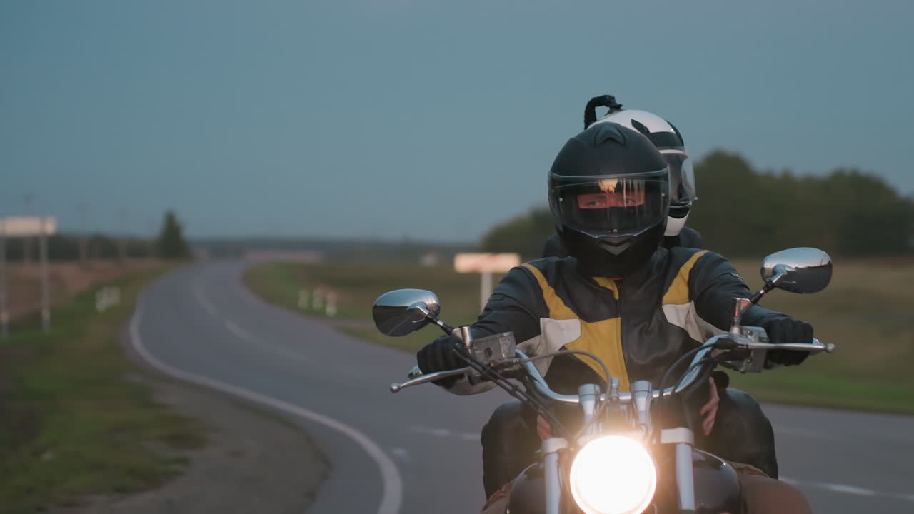 Man wearing black helmet and yellow-striped leather jacket rides motorcycle with woman seated behind him on curvy countryside road at dusk, surrounded by fields and trees under pale blue sky