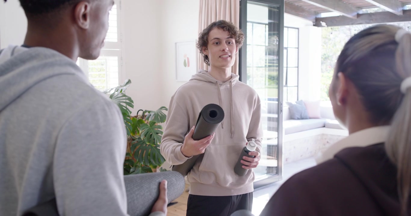 Amici di diversa etnia con felpe con cappuccio salutano un uomo al centro che entra in casa con un tappetino e una bottiglia, pronti per lo yoga