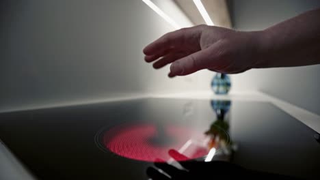 man placing a hand above a glowing red hot plate on the glass top of an electric hob with reflections and view along the counter to vase of tulips. wide shot